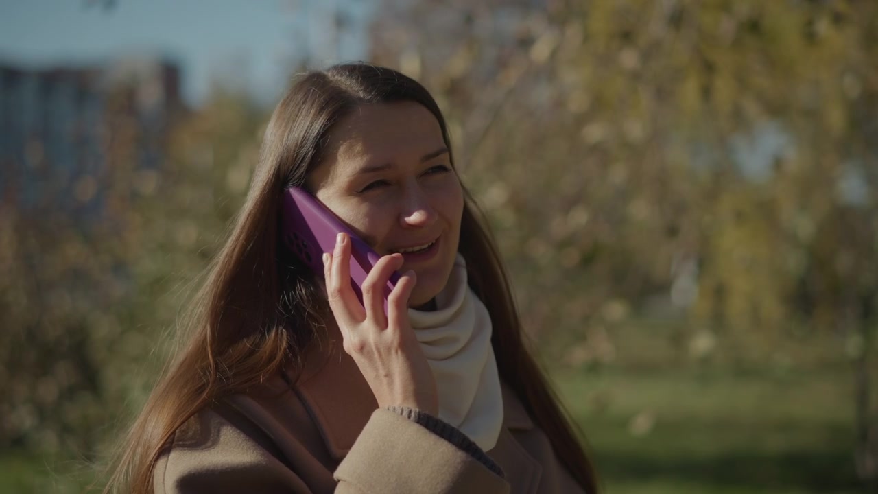 Primer plano de una mujer hablando por su teléfono morado al aire libre con luz natural. La mujer lleva un abrigo marrón y una bufanda blanca, con árboles y edificios visiblemente borrosos en el fondo. La escena es tranquila.