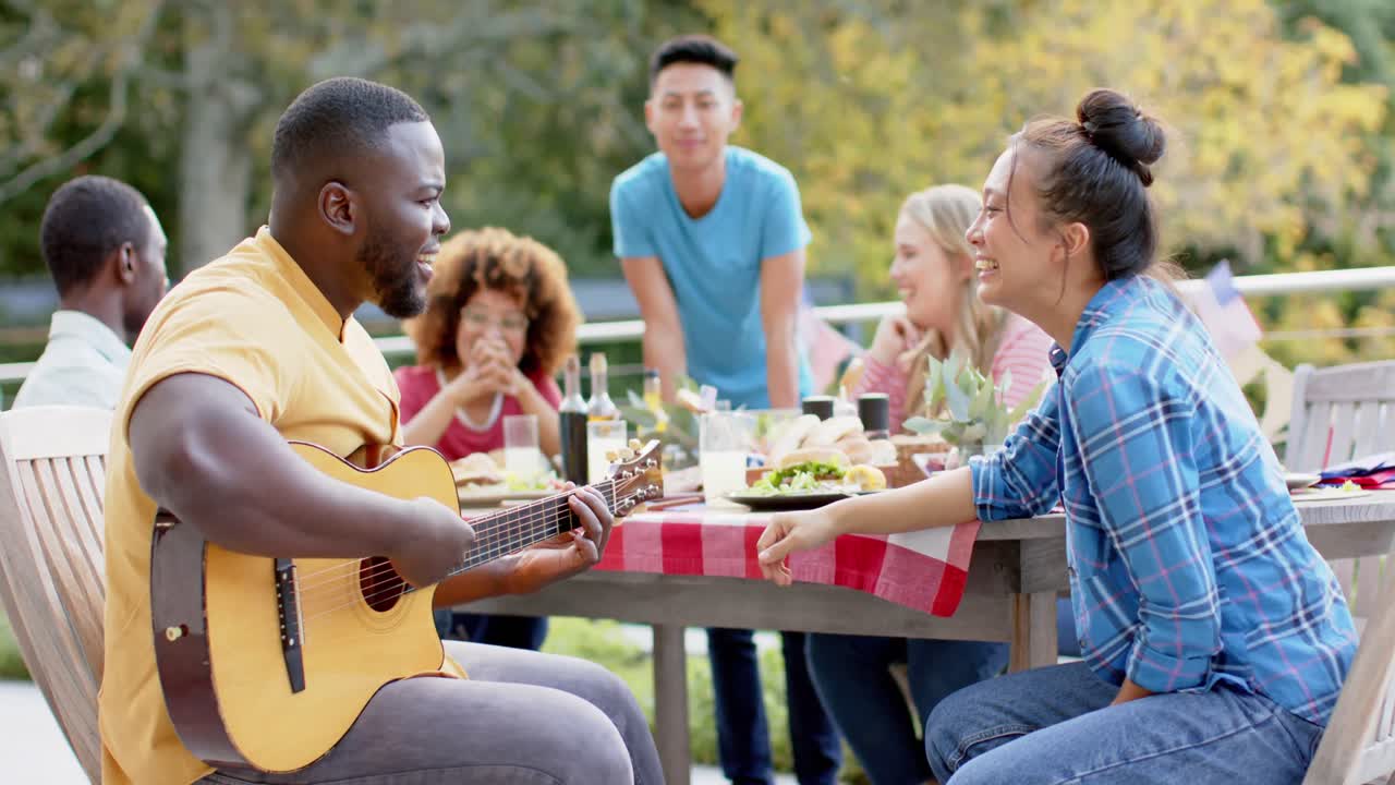 Uomo afroamericano che suona la chitarra con amici di diverse etnie a tavola in giardino, al rallentatore.