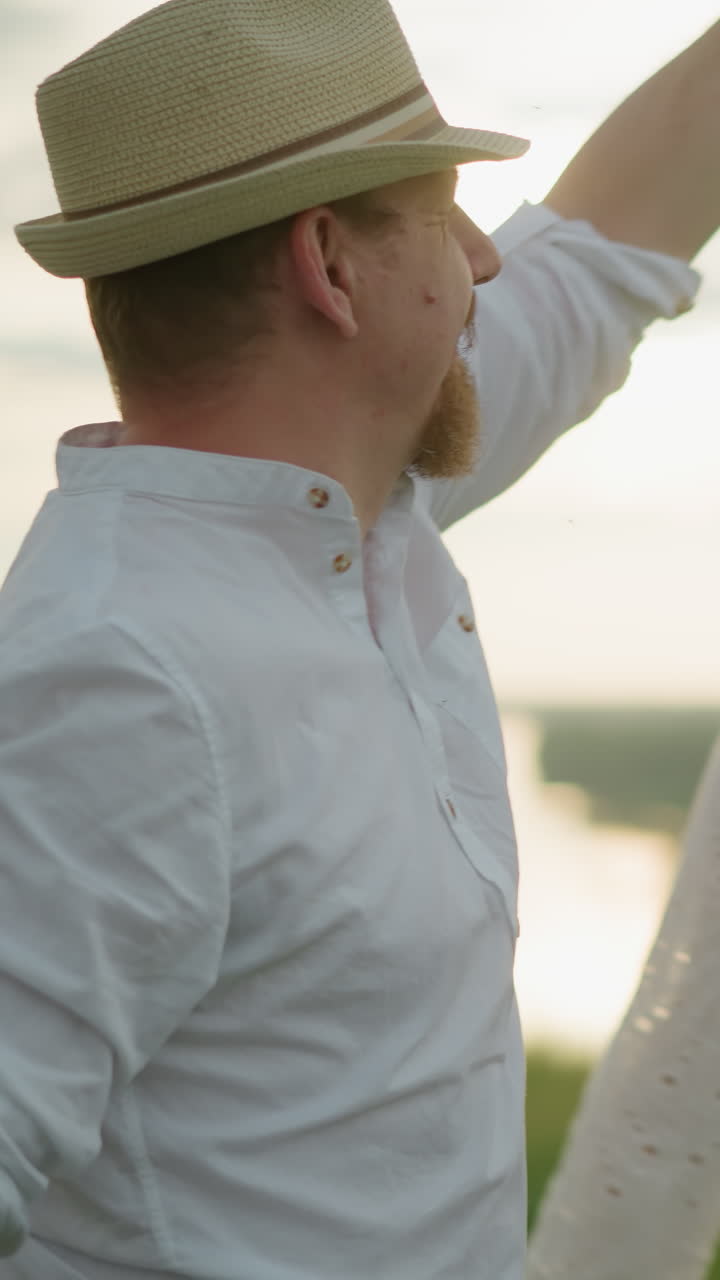 해가 지는 동안 평온한 호수 의 잔디 언덕에서 행복하게 아내를 회전시키는 남자의 클로즈업.  셔츠, 모자, 청바지를 입은 남자는 여자와 부드러운 순간을 공유합니다.