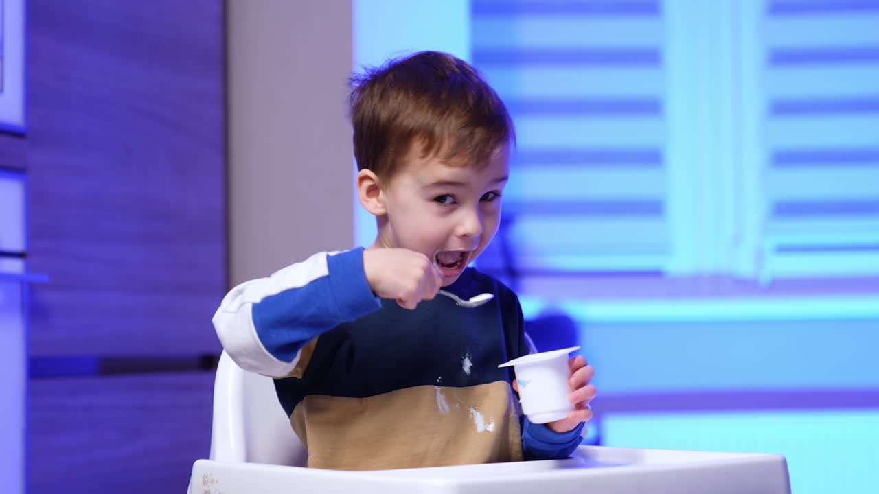 Little cute boy dirtied his sweater while eating yogurt. Lovely kid gets yogurt with a spoon from a cup.