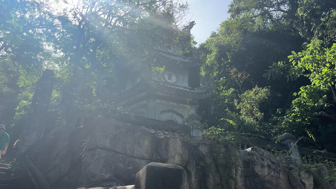 Stone temple building stands on rocky cliff at Marble Mountains near Da Nang, surrounded by dense green trees and sunlight filtering through foliage during daytime