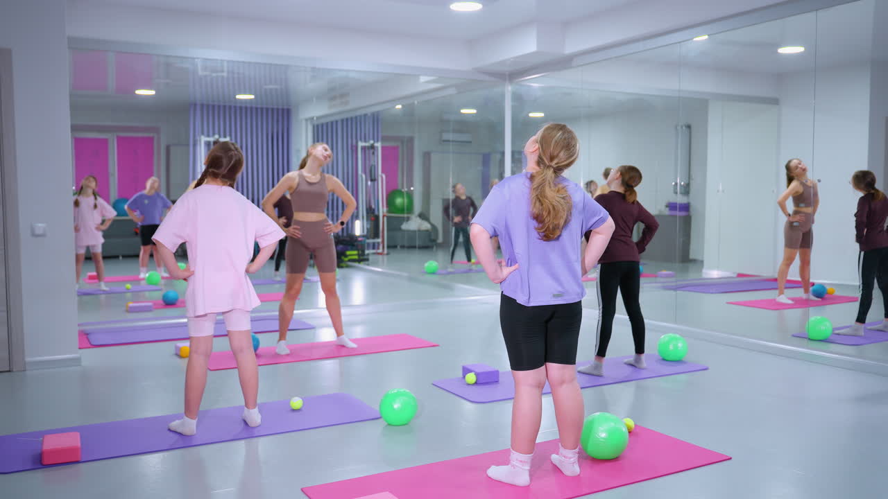 instructor de gimnasia conduce a tres mujeres a través de un ejercicio de torsión del cuello con las manos en la cintura, los participantes se paran en esteras de colores, rodeados de bolas de ejercicio y bloques, con espejos que reflejan la sesión