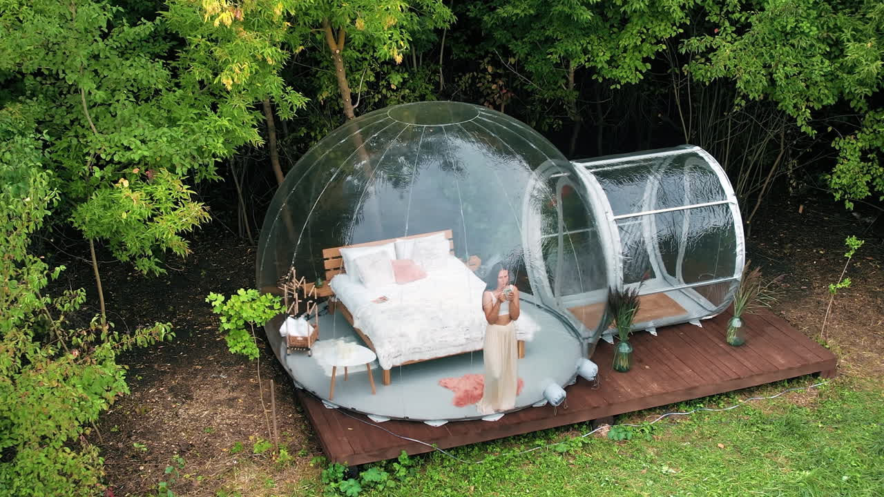 Gennemsigtigt bobletelt ved glamping, frodig skov rundt om og indvendigt. Kvinde med en kop, der kigger ud.