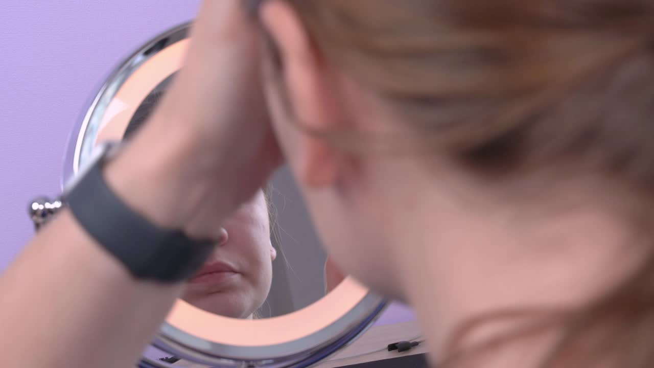 Young attractive woman applies the cream on the face looking at her reflection in round mirror, with rather strong movements. Shooting from the back. Lilac background, indoors.