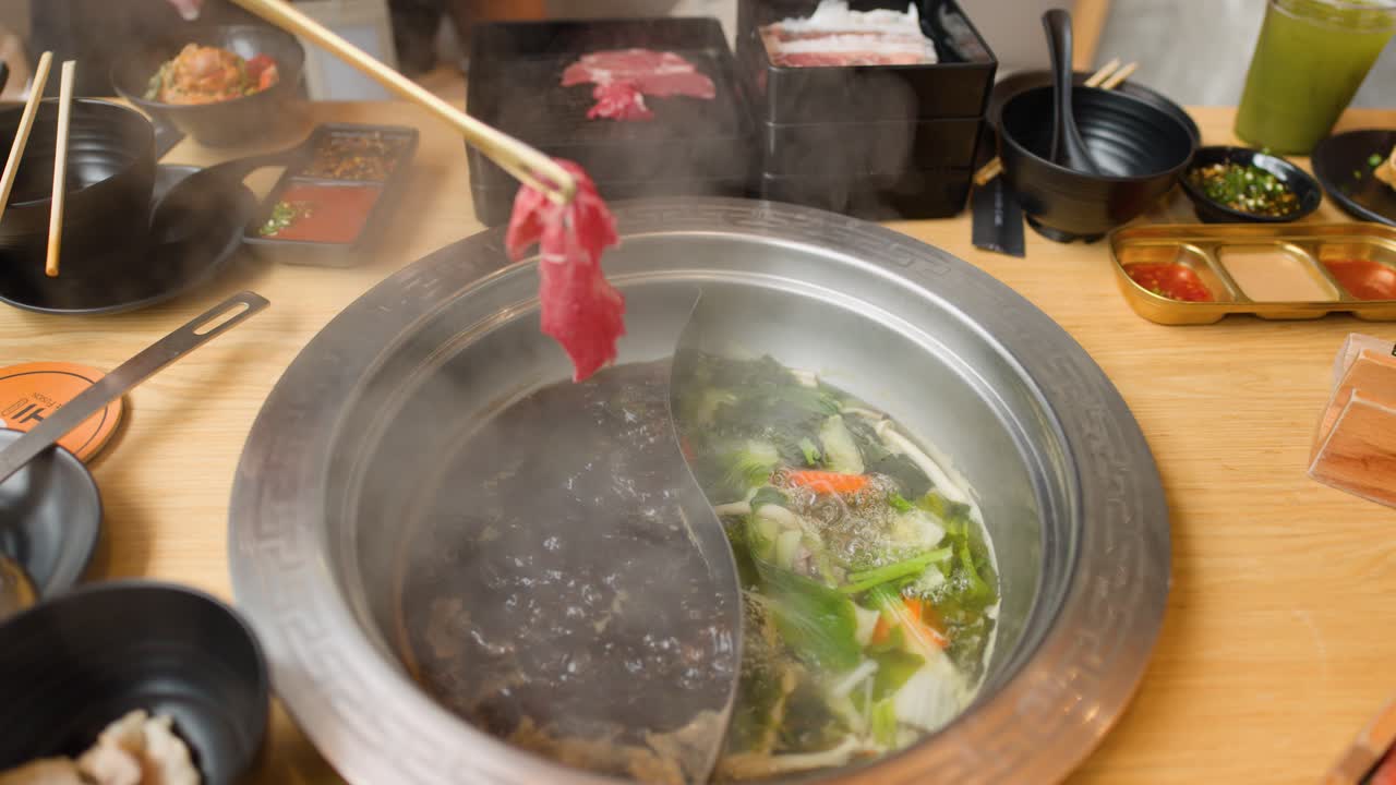 Hand cooks fresh beef slice in steaming dual broth hotpot, surrounded by vegetables and condiments