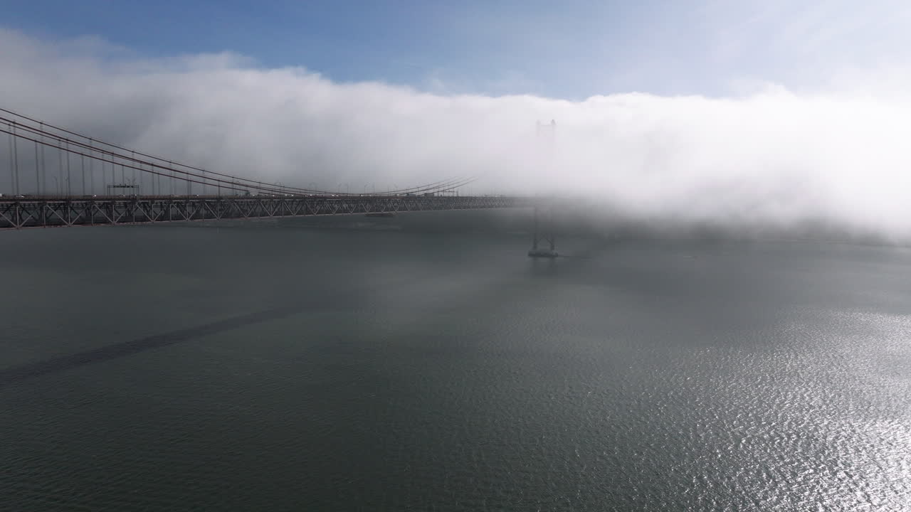 Aerial drone establishing shot towards the 25th April Bridge in Lisbon, Portugal, Europe. Sunny and bright with clouds, fog, mist, covering the bridge