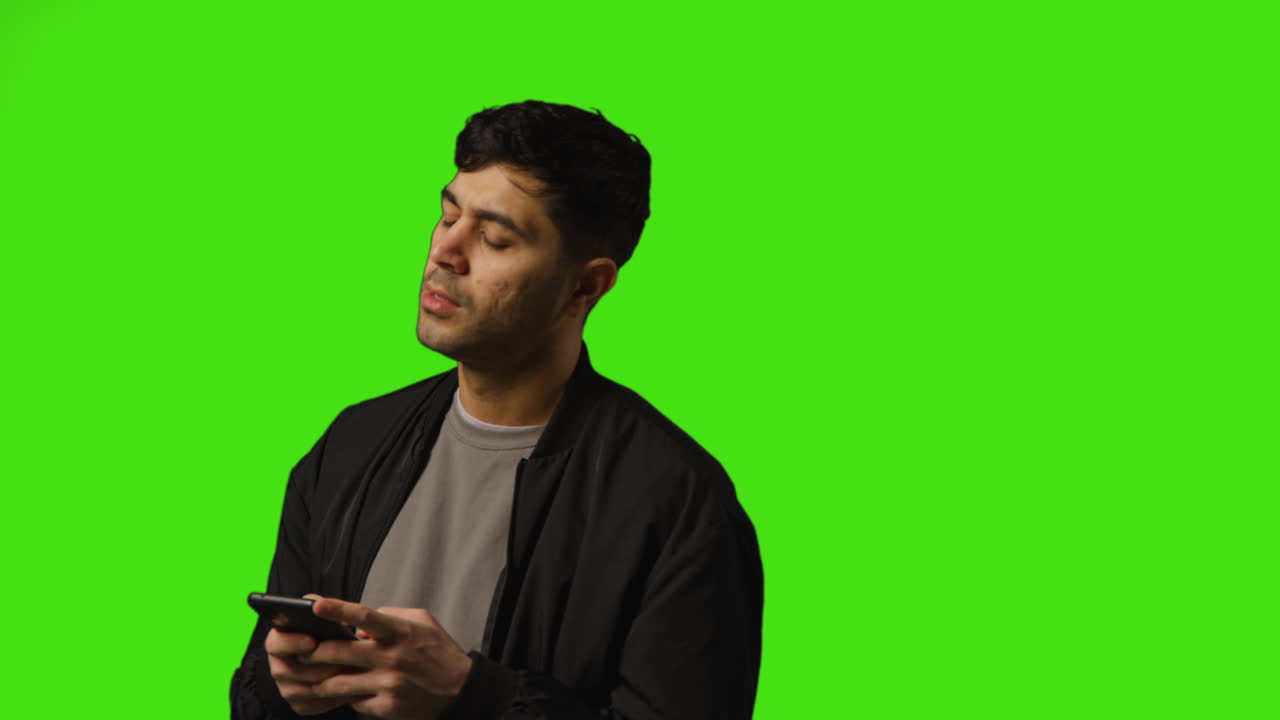Young Man Sending Text Message On Mobile Phone Standing Against Green Screen Background
