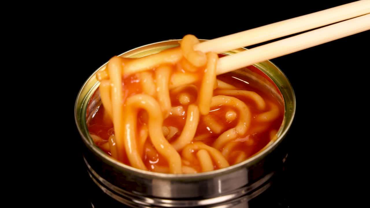 Chopsticks grasp and lift spaghetti noodles from a metal can filled with tomato sauce, set against a clean black background with bright, even lighting