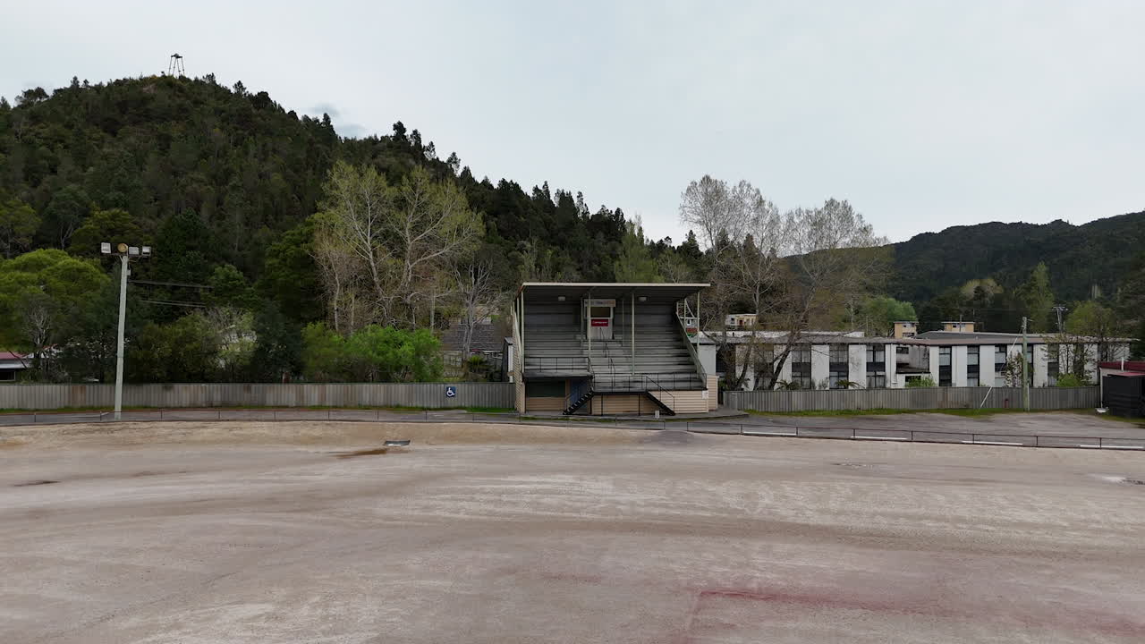 The Queenstown country football grandstand, showcasing its architecture set against a scenic backdrop of trees and rolling hills, with the field uniquely surfaced in gravel.