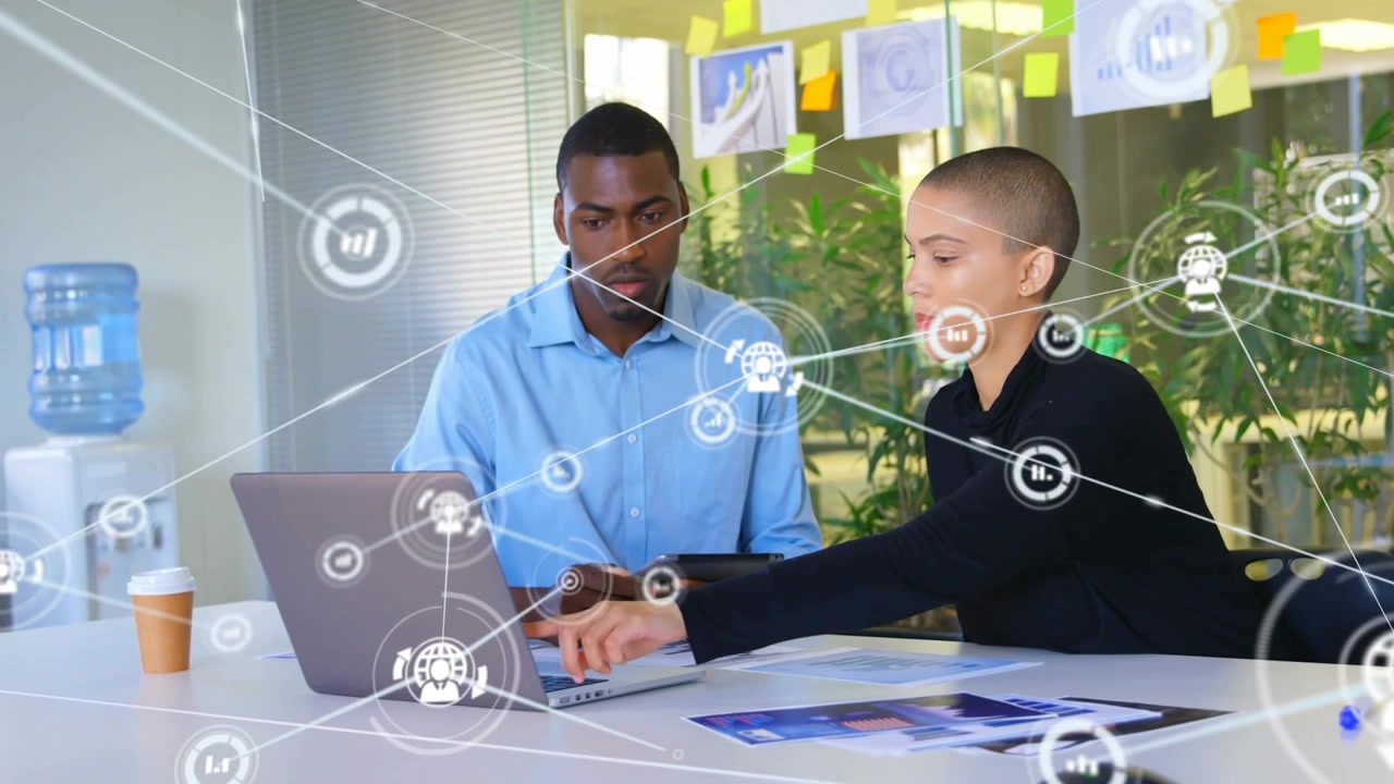 Woman pointing at laptop trackpad while man watching tablet as technology icons appearing for data