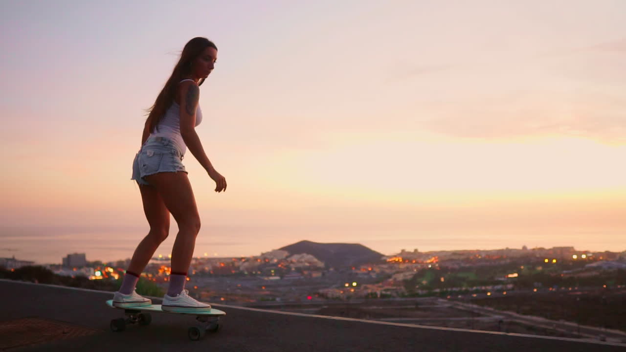 Amid the tranquil sunset, a young and fashionable skateboarder gracefully rides her board in shorts along a mountain road, the mountains' view captured in slow motion