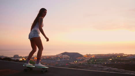 Amid-the-tranquil-sunset,-a-young-and-fashionable-skateboarder-gracefully-rides-her-board-in-shorts-along-a-mountain-road,-the-mountains'-view-captured-in-slow-motion