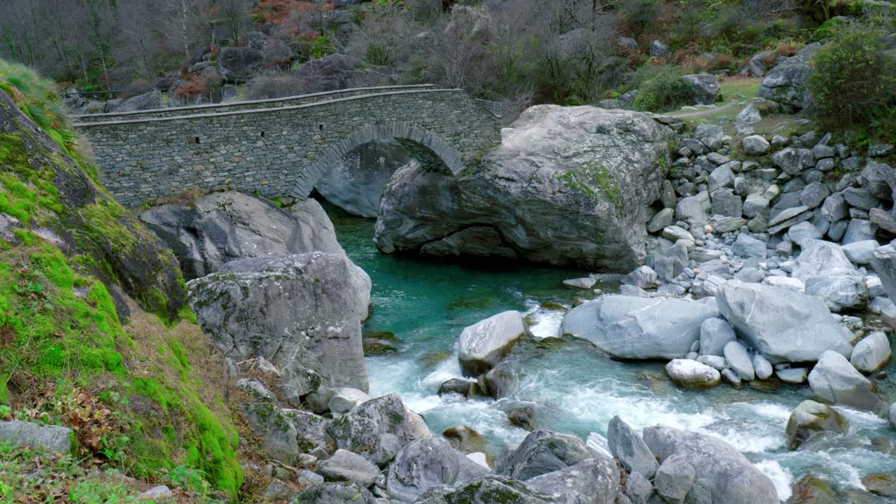 el dron se mueve lentamente desde abajo hacia arriba, mostrando en su marco un puente, aguas glaciales que fluyen, y las rocas a lo largo de las orillas del río maggia, en el pueblo de cavergno en suiza.