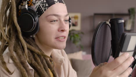 close up view of woman with dreadlocks recording a podcast wearing headphones, talking into a microphone and reading on smartphone sitting at a desk