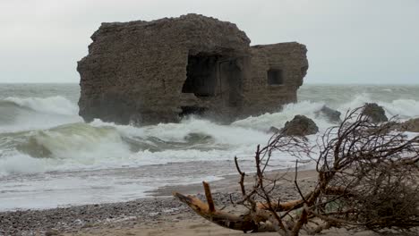 big stormy waves breaking against abandoned seaside fortification building ruins at karosta northern forts in liepaja, baltic sea, wave splash, pine branches in front, overcast day, medium shot