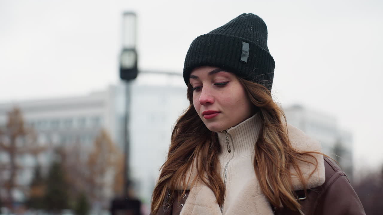 Close shot of lady in black cap brown jacket walking with thoughtful expression against blurred city background under cloudy autumn sky showing emotion in serene peaceful atmosphere