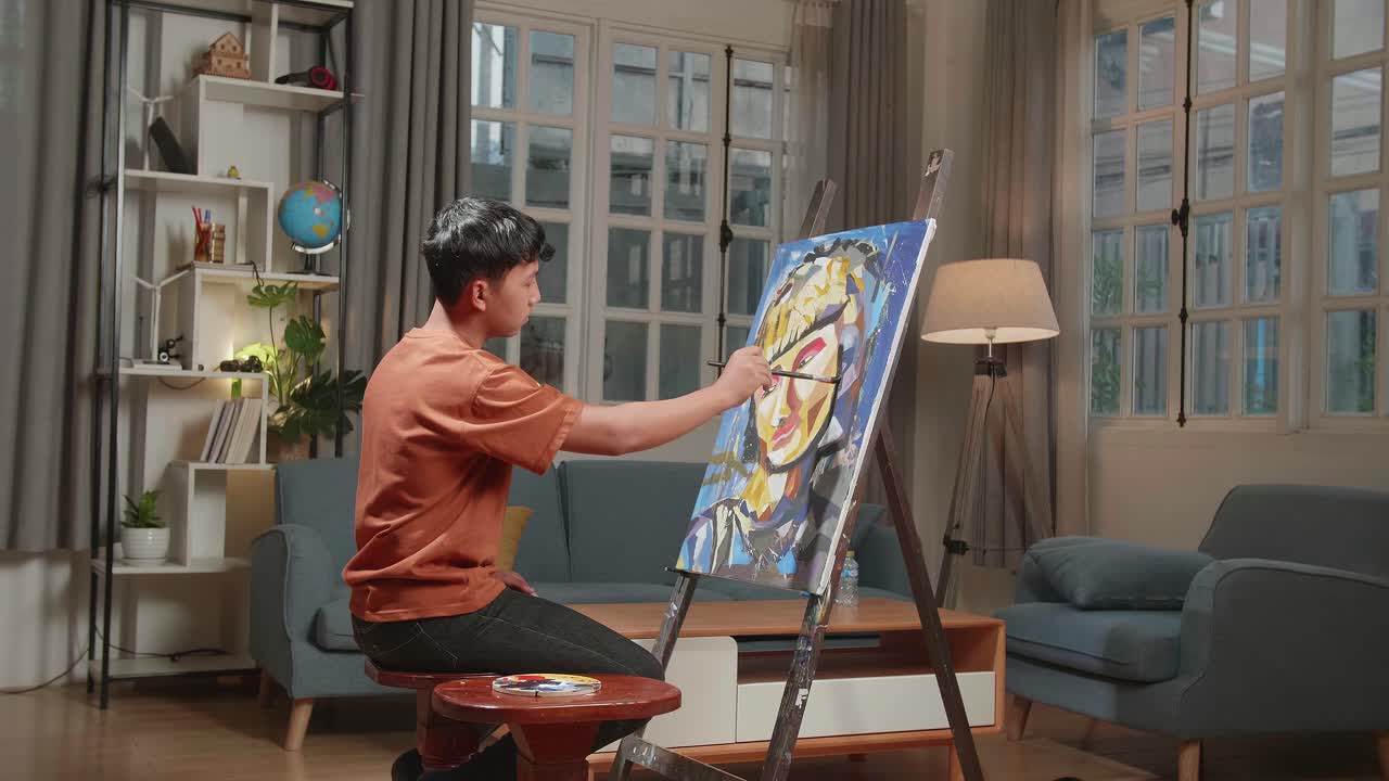 Asian Artist Boy Sitting On Round Wooden Chair Without Backrest Holding Paintbrush Mixed Colour And Painting A Girl'S Hair On The Canvas