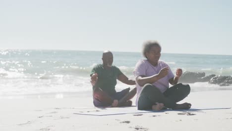 happy senior african american couple doing yoga and meditating at beach, in slow motion