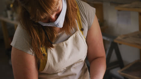 Female-potter-wearing-face-mask-and-apron-creating-pottery-on-potters-wheel-at-pottery-studio