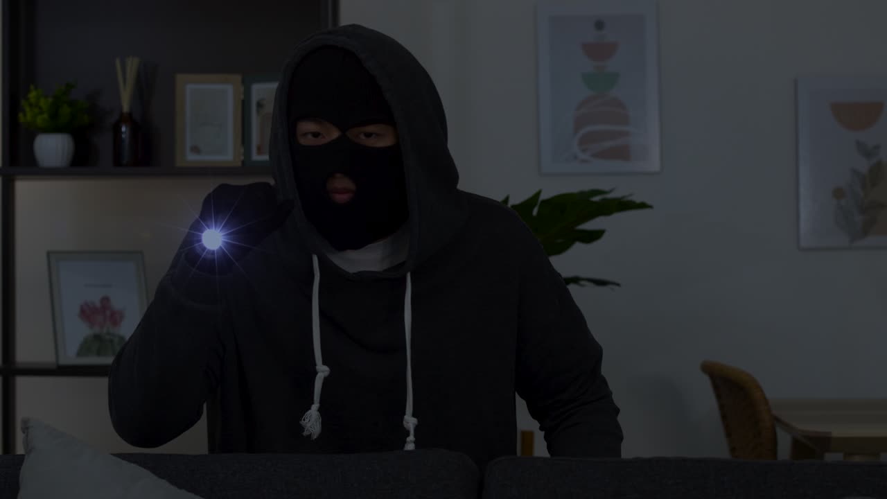 Ein maskierter Eindringling in einem dunklen Kapuzenpulli durchsucht ein Wohnzimmer mit einer hellen Taschenlampe.