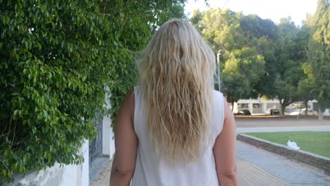 Slow-motion-view-of-a-blonde-woman-walking-down-a-tree-lined-residential-street,-rear-view