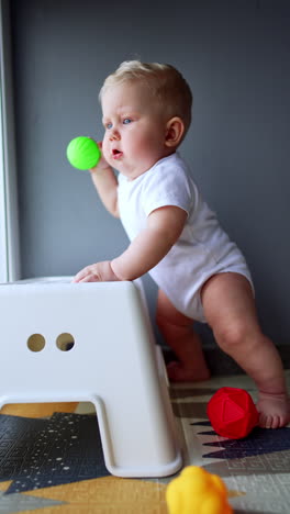 adorable plump blond boy in white bodysuit stands near the chair. lovely child holds a ball in his hand. vertical video.