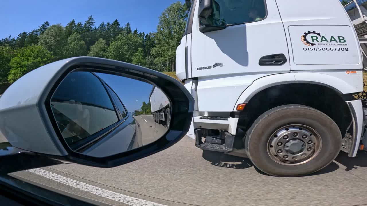 Side mirror view of highway drive past forested roadside and passing cargo truck in summer