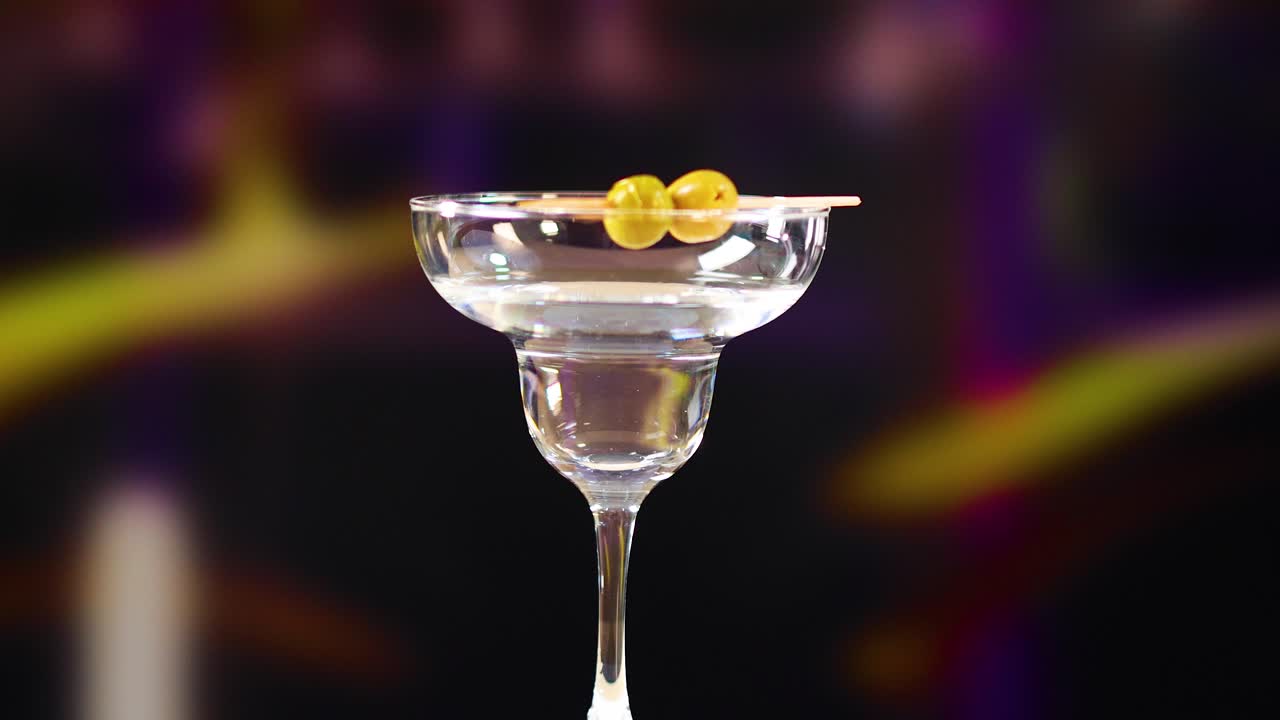 A hand places olives in a martini glass, set against a colorful, blurred background with soft lighting