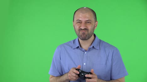 portrait of a middle aged men playing a video game using a wireless controller with joy and rejoicing in victory. balding male with beard in blue shirt posing on green screen in the studio. close up