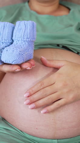 leaned back pregnant woman on the couch stroking her big belly with one hand. in her other hand she's holding baby booties and shoving them closer to the camera while camera moves back. vertical video