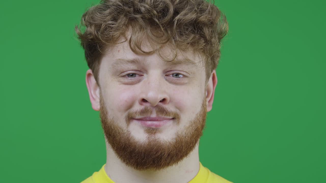 портрет мужчины с серыми глазами на зеленом экране в студии. рыжеволосый мужчина с кудрявыми волосами и веснушками смотрит прямо в камеру и улыбается. близко. медленное движение готово 59.97fps