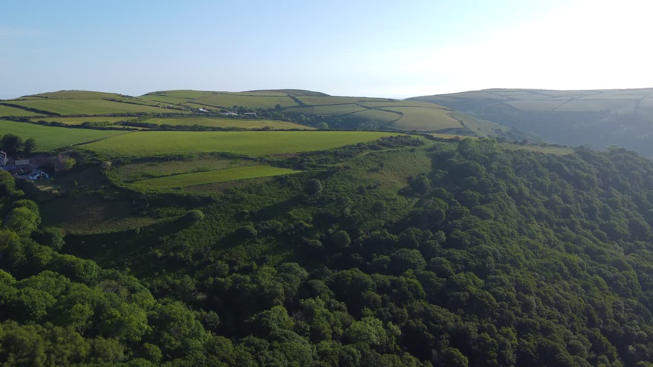 Sprawling British Countryside Fields Seen from the Air with Dense Dark Forests Lining Valley Side with Early Morning Sun Flare with Blue Sky
