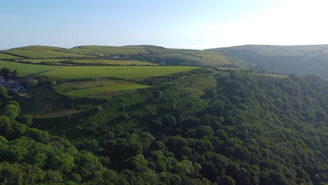 sprawling british countryside fields seen from the air with dense dark forests lining valley side with early morning sun flare with blue sky