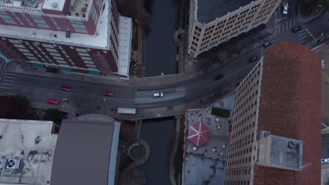 A drone captures a top-down view of a river winding through the city, with bridges and reflections of skyscrapers on the water.