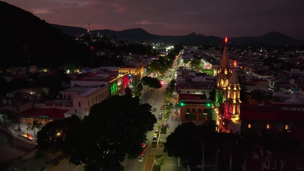 Dolly in aerial at night overlooking the Saint Francis Parish, Town Hall building, Chapala city center and the Francisco Madero avenue decorated for Mexico Independence Day