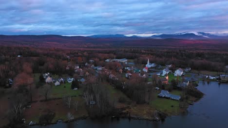 Luftaufnahmen,-Die-Bei-Sonnenaufgang-Im-Spätherbst-über-Den-See-Fliegen,-In-Richtung-Einer-Kleinen-Ländlichen-Gemeinde-Mit-Nebligen-Bergen-In-Der-Ferne