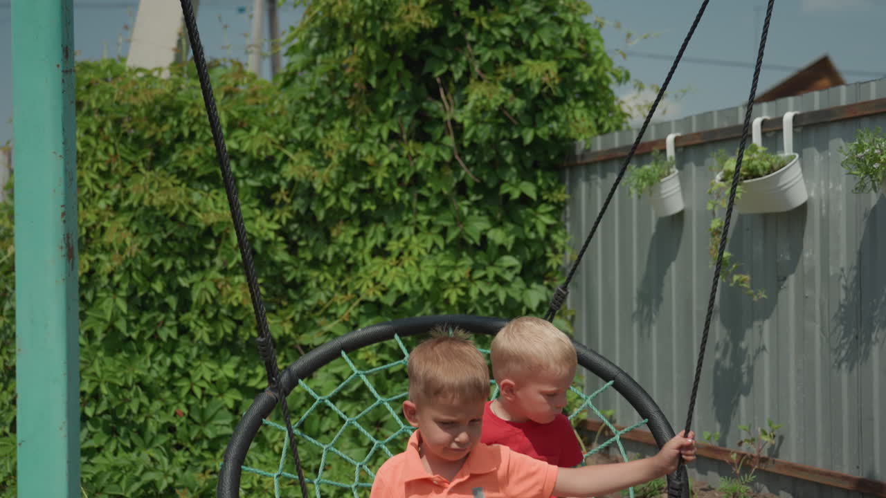 Caucasian Boys On Nest Swing In Backyard, Smiling And Holding Ropes During Sunny Summer Afternoon, Leafy Fence And Punching Bag In Background, Candid CloseUp Of Playful Sibling Bonding And Quiet Joy