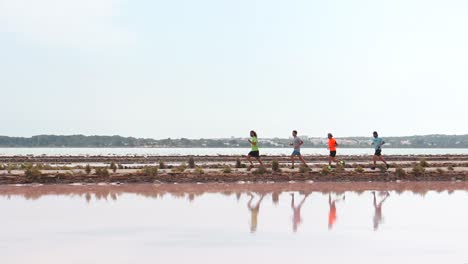 smooth pan shot from right ro left of four men running in front of a lake in formentera. you can see its reflections in the water, and behind some beautiful houses