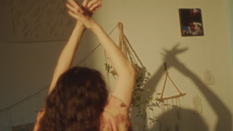 relaxed girl dancing with raised arms, casting shadows on sunlit wall at home