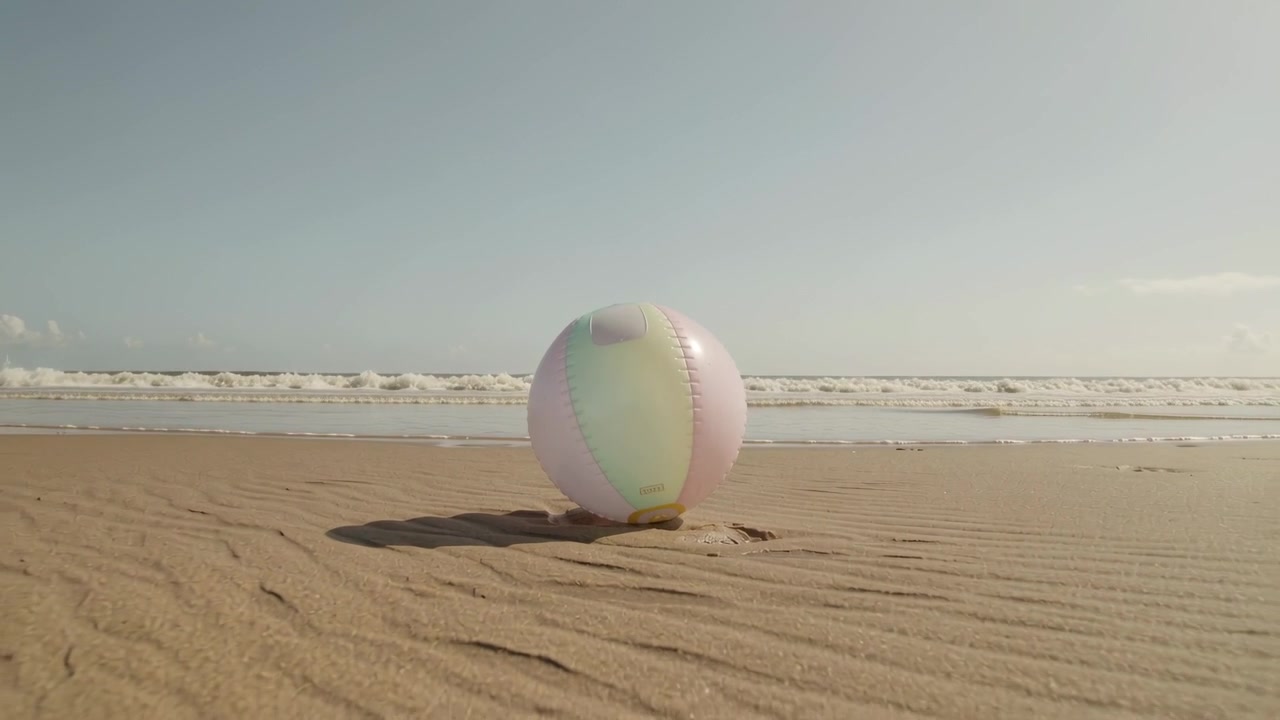 Pulling back, camera revealing pastel beach ball resting on rippled sand dip near shoreline, waves
