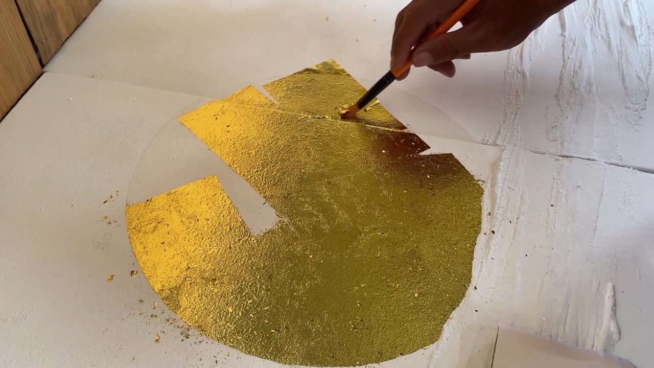 Handgehaltene 4K-Nahaufnahme von Händen, die schimmerndes Blattgold auf eine Leinwand auftragen. Selektiver Fokus fängt den künstlerischen Prozess der Schaffung luxuriöser strukturierter Wandkunst ein