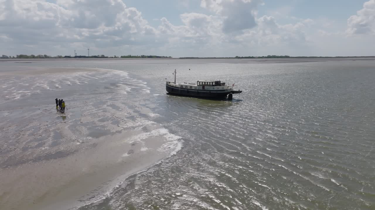 Group of people standing on exposed mudflat at low tide with a classic vessel in the background; aerial daylight view. Wadden Sea