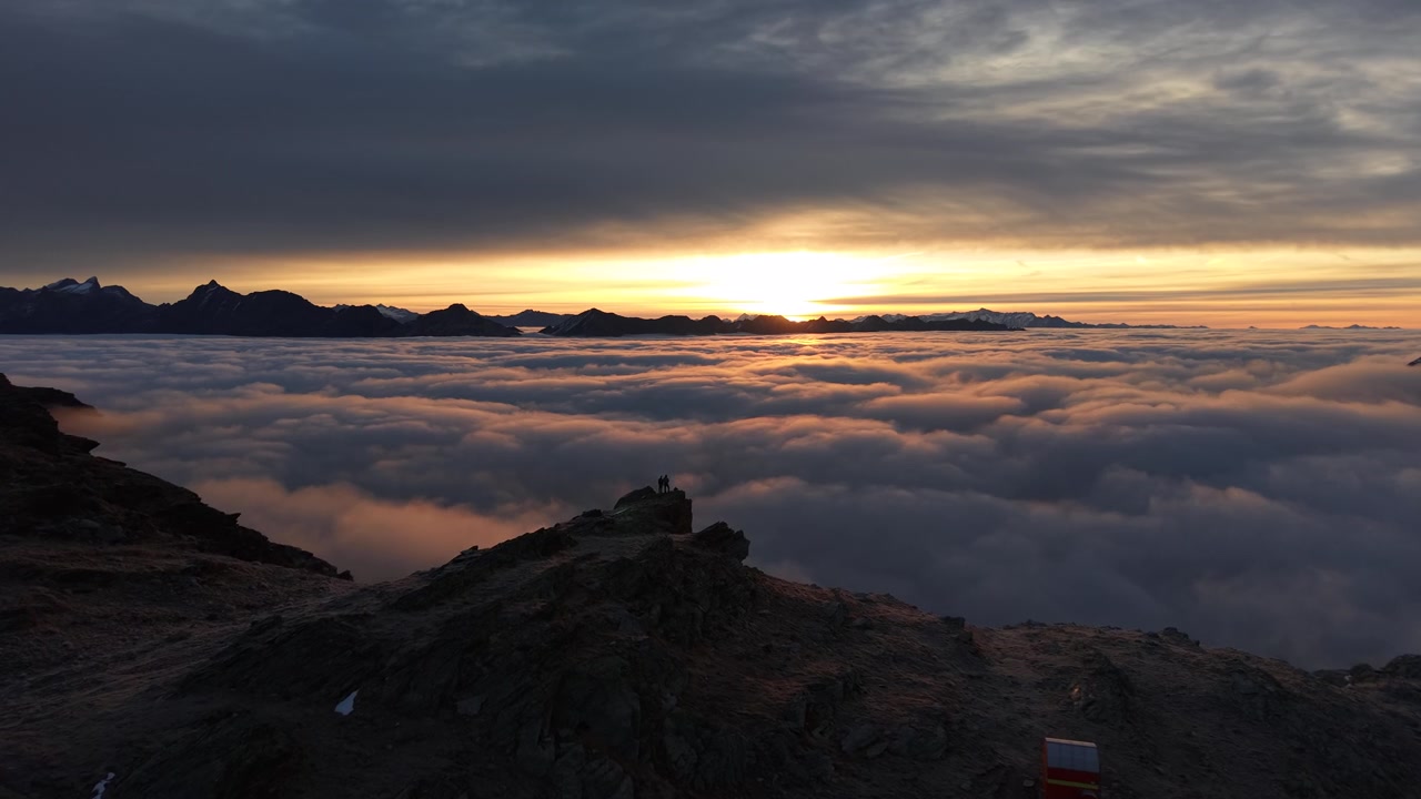 Sorvolo aereo sopra escursionisti che si godono il bagliore dell'alba sul mare di nuvole nelle Alpi Svizzere con una vista panoramica delle montagne e del cielo colorato.