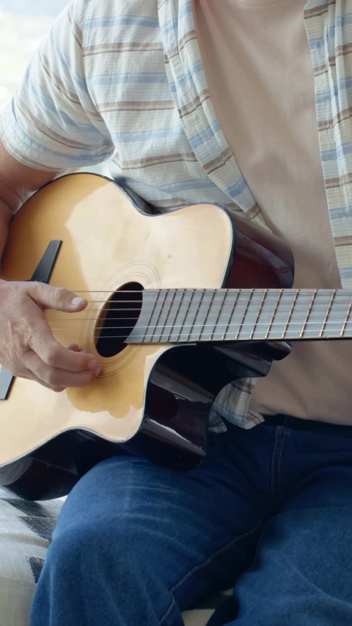 Video vertical: Hombre sentándose en una silla, rasgueando la guitarra y practicando una canción en el porche, con camisa a rayas.