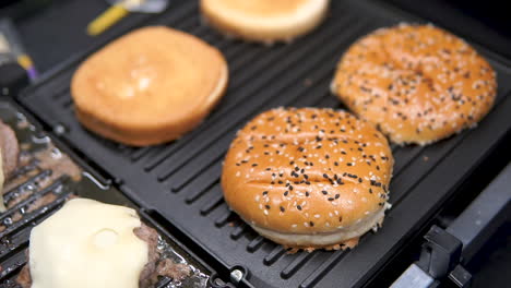 Rindfleischburger-Mit-Gelbem-Käse-Werden-Auf-Einer-Schwarzen-Grillplatte-Gebraten,-Während-Die-Brötchen-Für-Ein-Köstliches-Hausgemachtes-Burgerbrötchen-Vorgewärmt-Werden