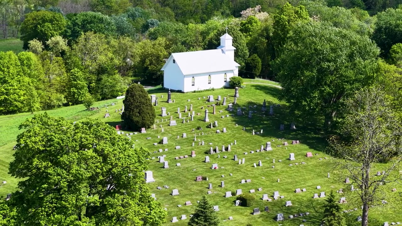 Vista aerea di una chiesa di campagna bianca che si affaccia su ordinate file di frutteti all'interno di una lussureggiante campagna boscosa.