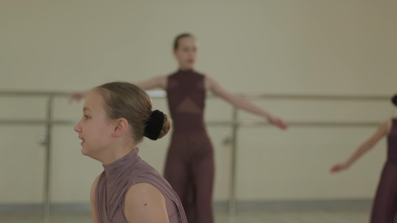 Skupina mladých studentů baletu v černém tanečním oblečení procvičuje pozice v prostorném baletním studiu s dřevěnou podlahou a nástěnnými tyčemi. Soustředěné výrazy a synchronizované pohyby