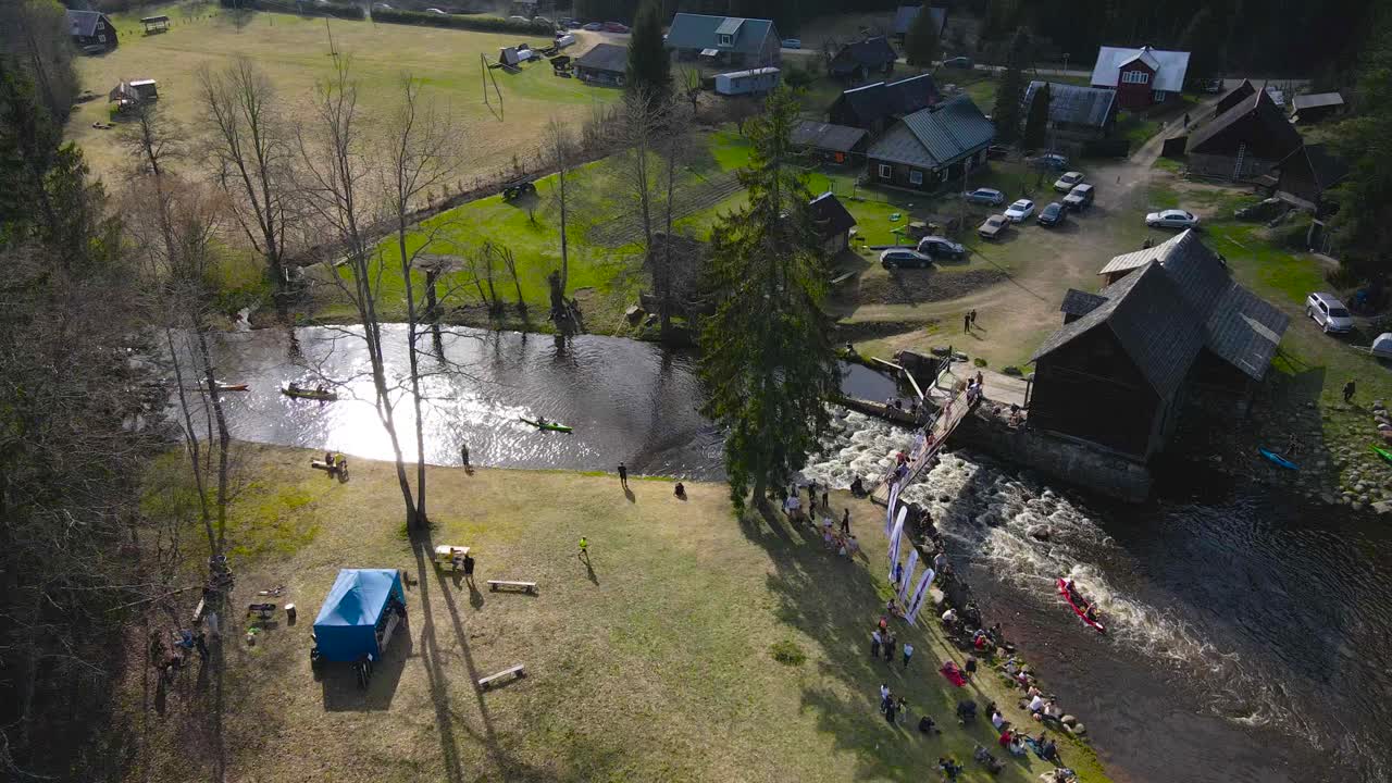 Gorgeous aerial drone footage panning over colorful kayak canoes and rafts that are going through dark narrow river rapids with foamy water under a bridge during a sunny day at Võhandu marathon.