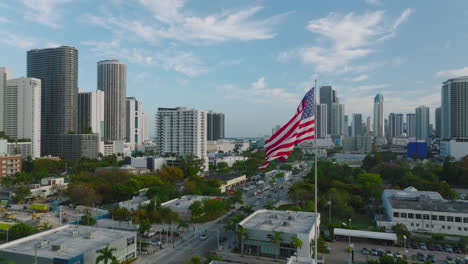 Orbit-shot-around-US-flag-waving-on-pole-on-top-of-house.-Panoramic-view-of-high-rise-downtown-buildings.-Miami,-USA