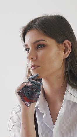 thoughtful long haired young woman artist with grey paint on palm in casual clothes looks at canvas on wooden easel in light studio slow motion closeup